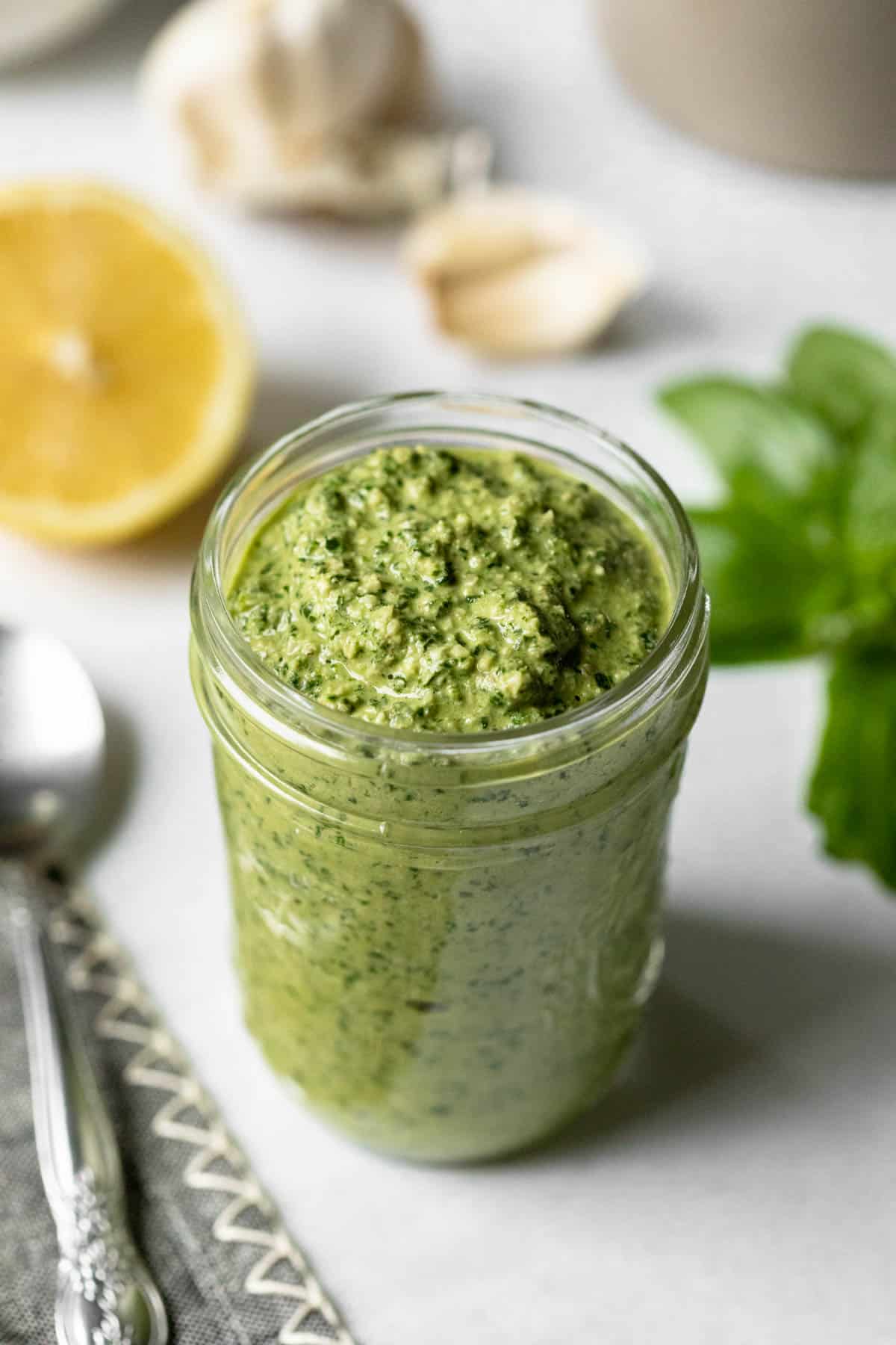 bright green fresh vegan pesto in a small glass jar with lemon, garlic, and basil in background.