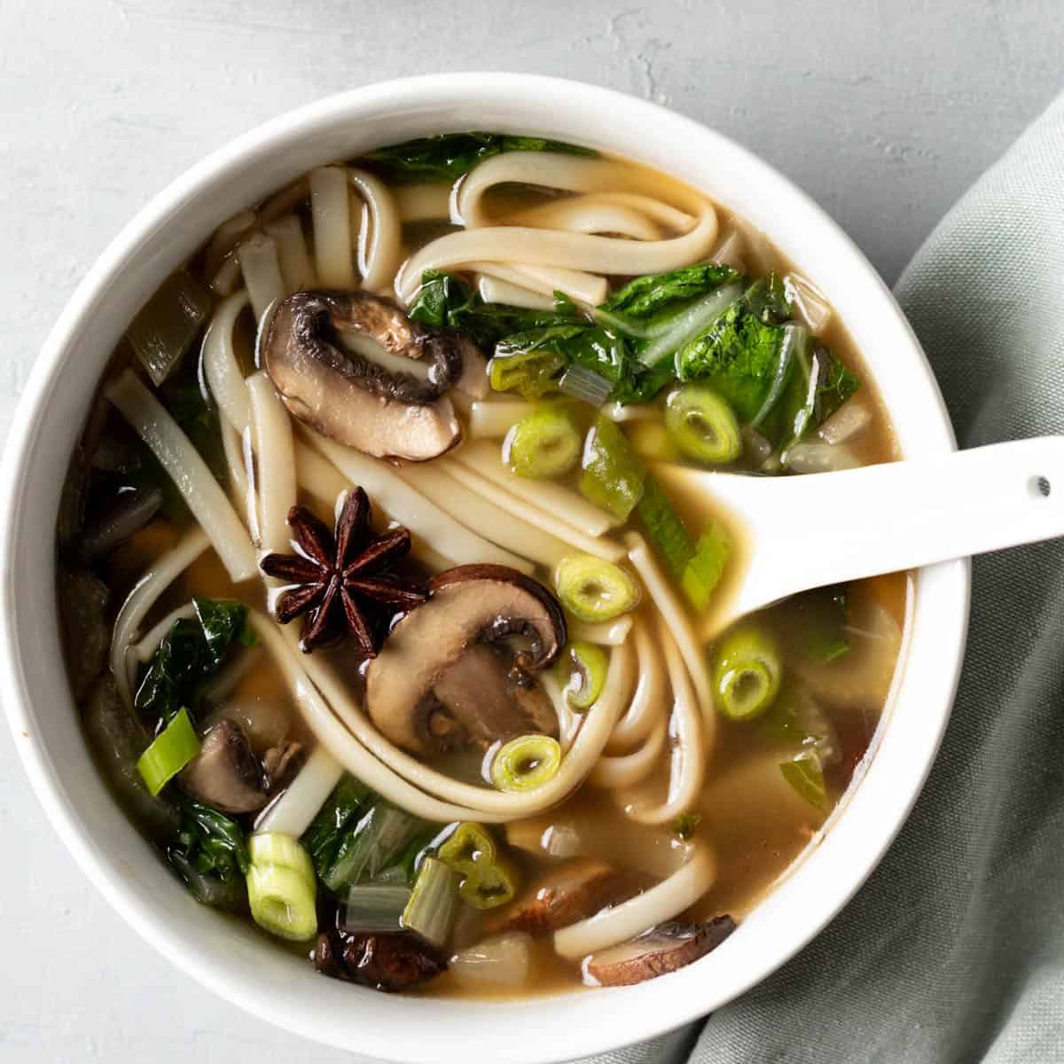 A bowl of bok choy noodle soup with mushrooms and star anise with a white spoon resting in the bowl.
