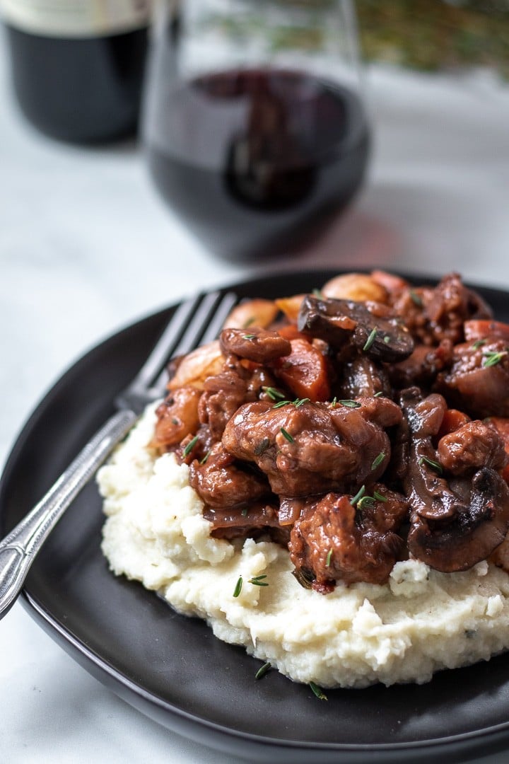Seitan Bourguignon With Rosemary Cauliflower Mash on a black plate with a glass of red wine in background