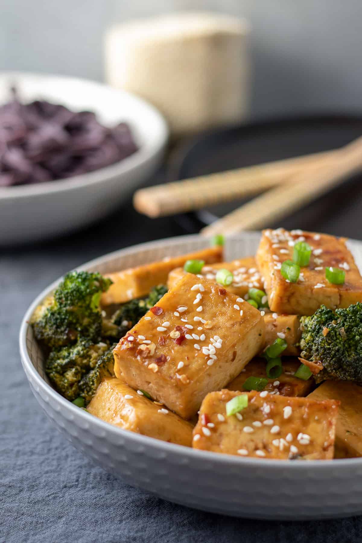 Sticky Sesame Tofu With Broccoli in a gray bowl with noodles and chop sticks in background.