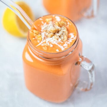 orange colored carrot smoothie in a glass mug topped with coconut and walnuts.