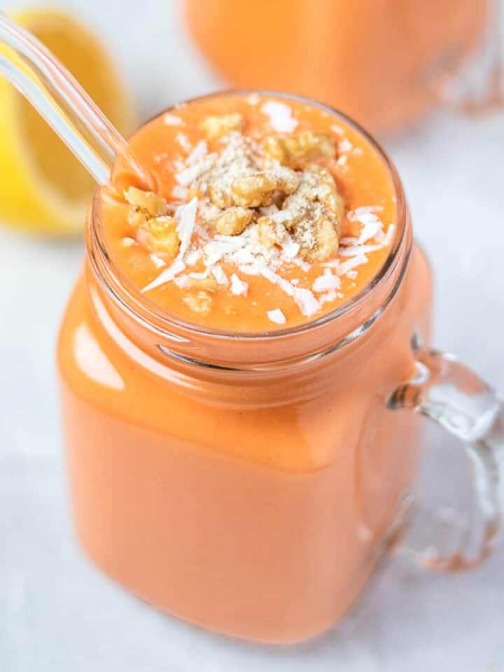 orange colored carrot smoothie in a glass mug topped with coconut and walnuts.