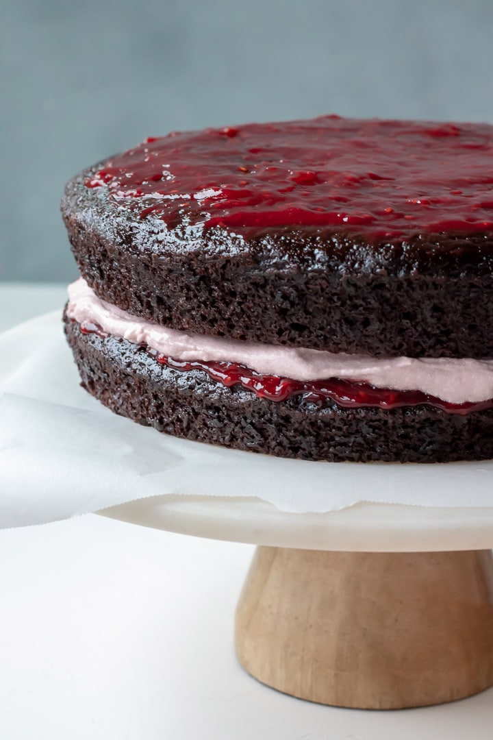 layers of chocolate cake spread with raspberry jam, then raspberry frosting, sitting on a cake stand.