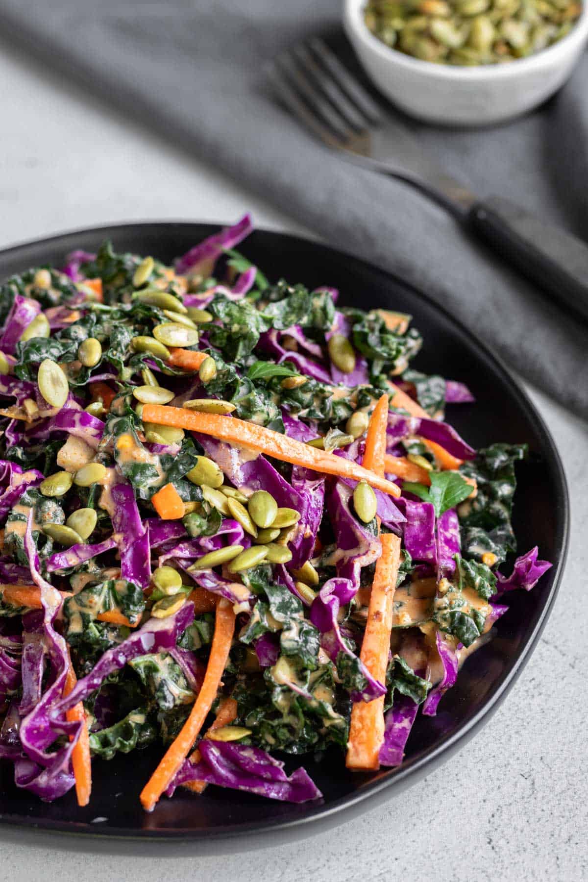 Kale slaw with carrot, red cabbage, and colorful harissa dressing on a black plate.