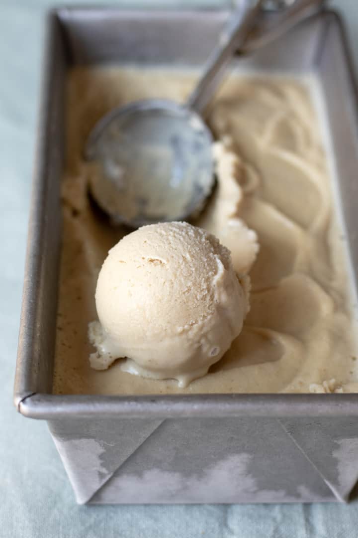 an ice cream scoop resting in a frosty loaf pan filled with vegan vanilla ice cream.