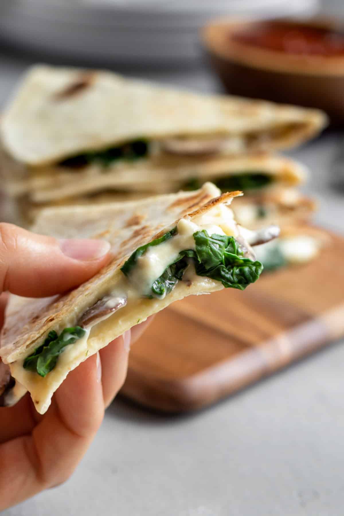 hand holding a triangle of vegan quesadilla showing the melted cheese and warm spinach.