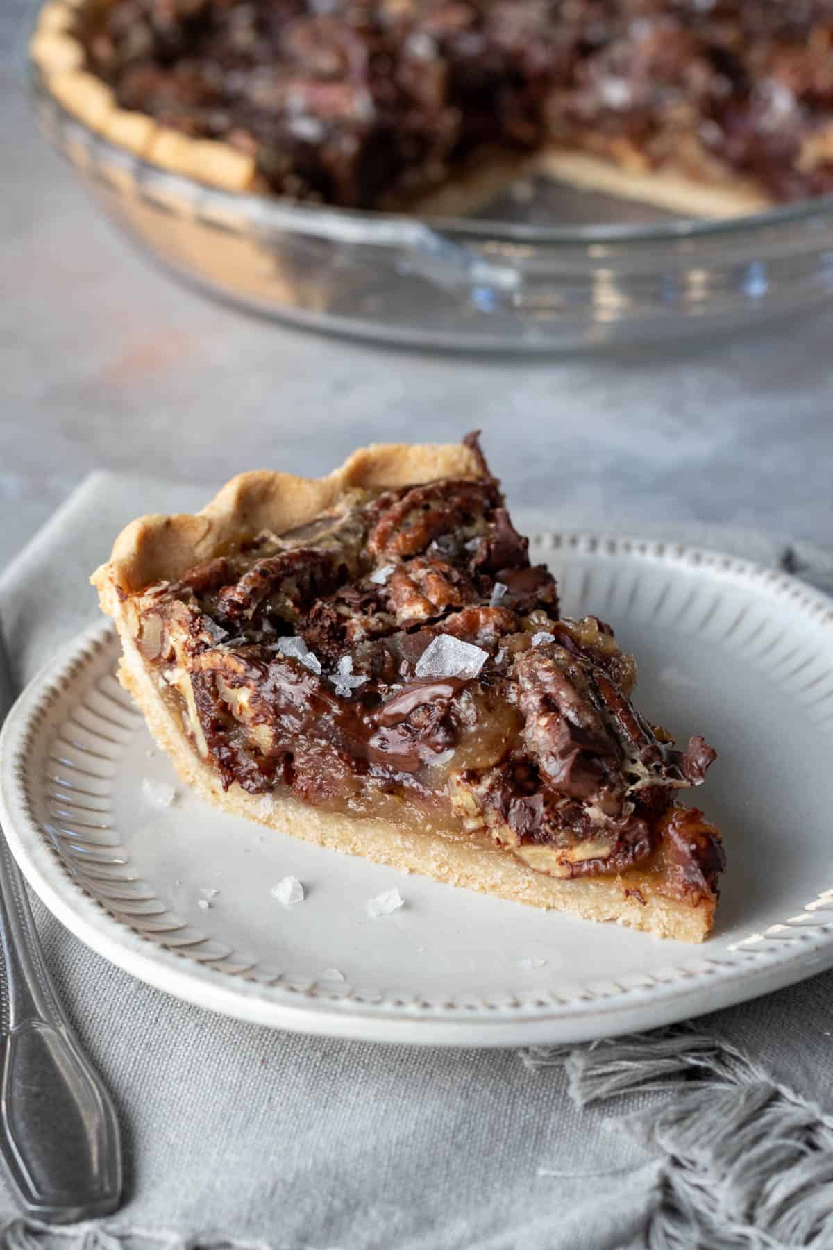 a slice of salted chocolate pecan pie on a plate with the whole pie in background.