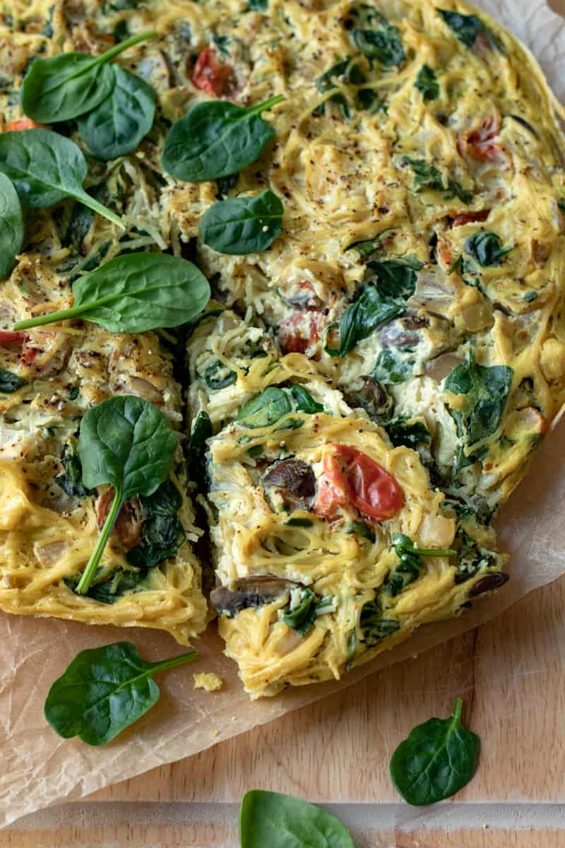 pasta frittata on a cutting board topped with a handful of fresh spinach