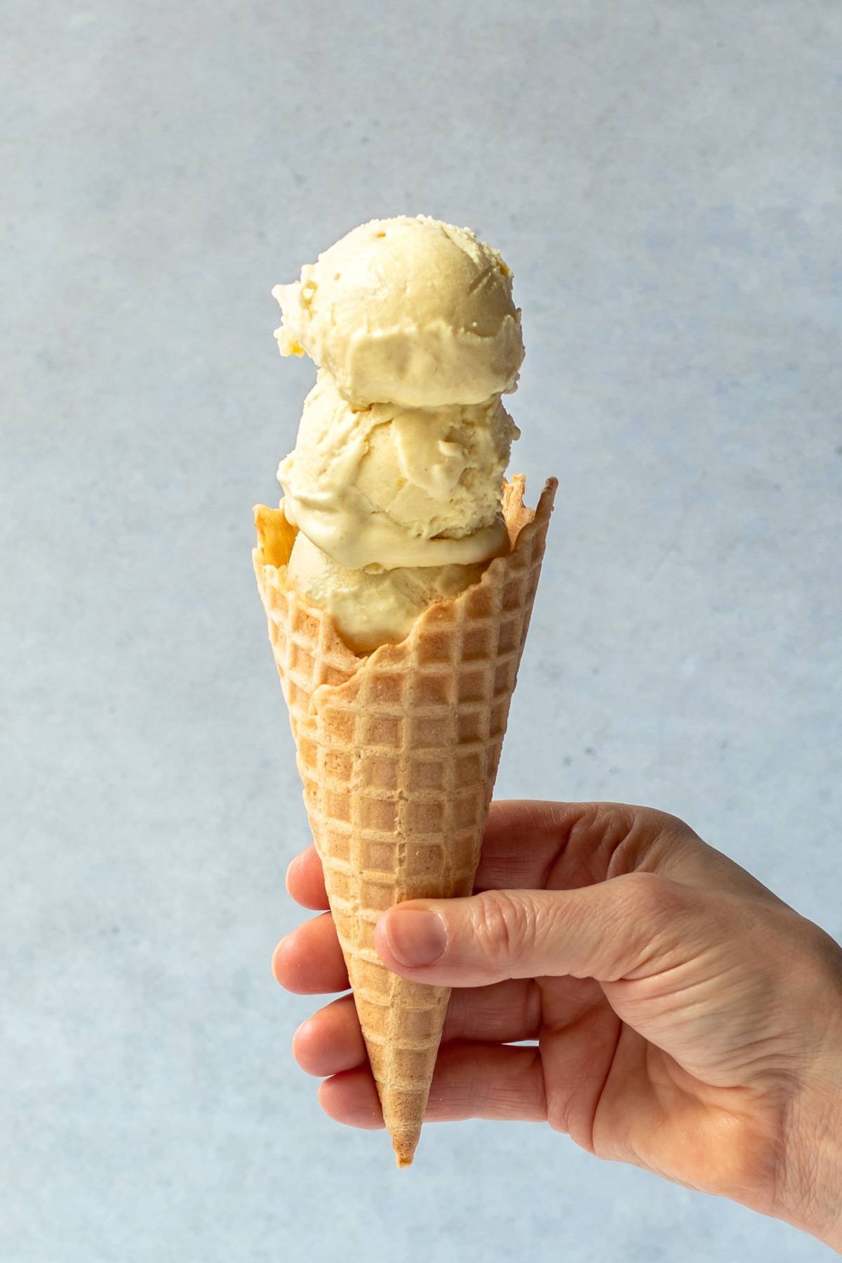 hand holding a waffle cone filled with 3 scoops of creamy lemon ice cream.