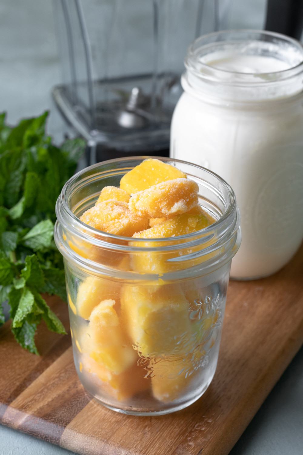 jar of cashew yogurt and jar of mango chunks with mint and blender in background