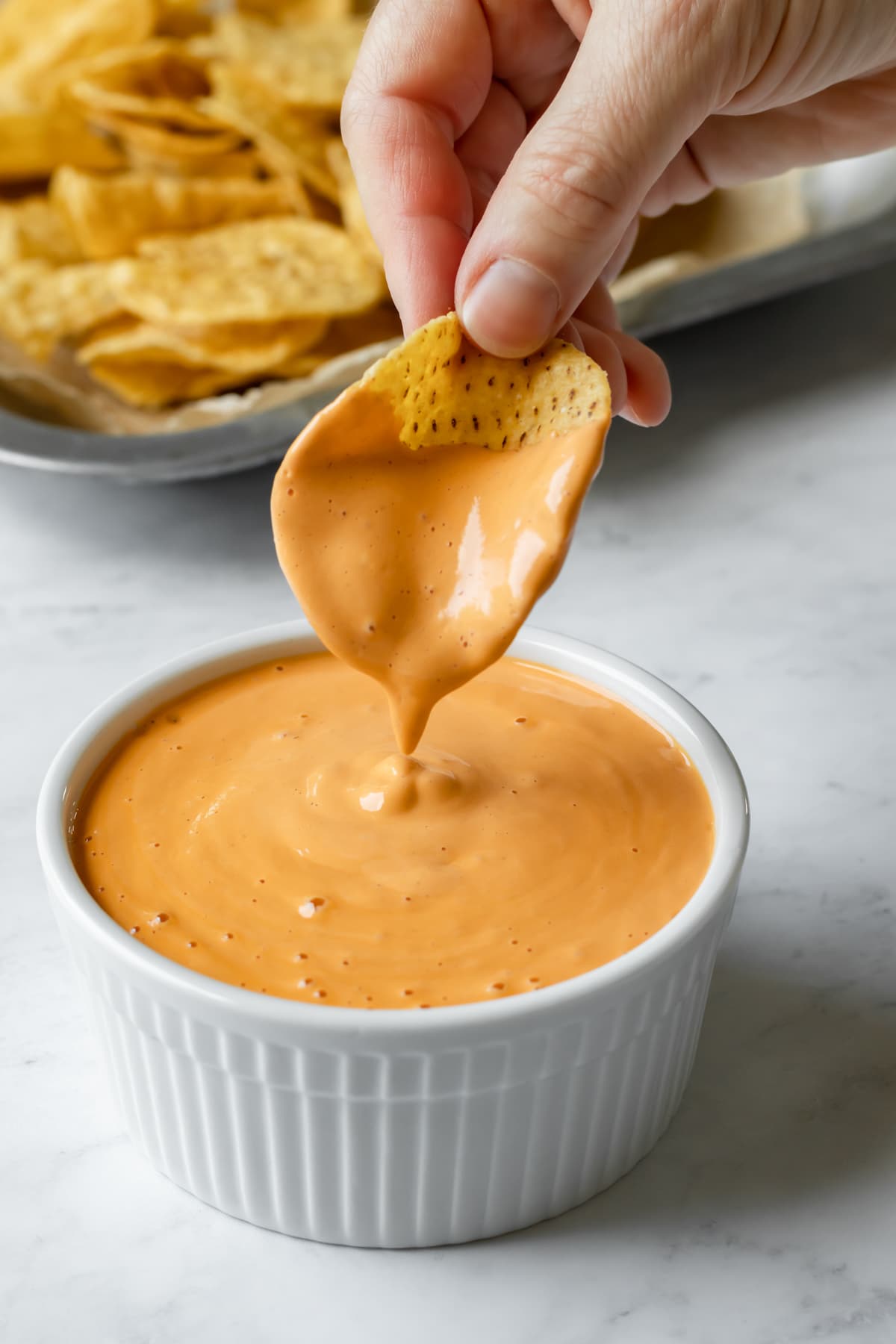 A hand dipping a tortilla chip in a small bowl of cheesy sauce.