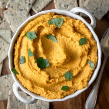 Bright orange carrot hummus in a small white dish surrounded by crackers.