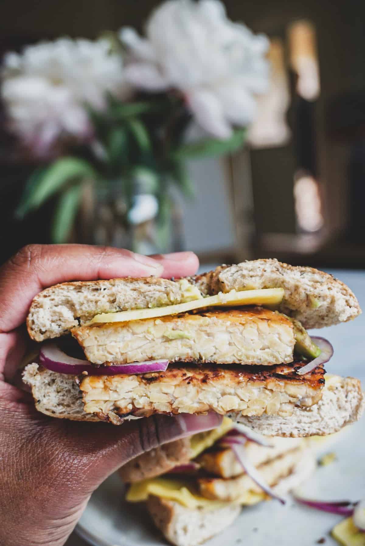 hand holding a bagel sandwich showing thick slices of tempeh.
