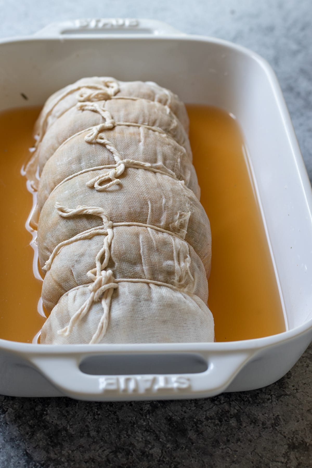 seitan wrapped in cheese cloth, tied with twine, resting in broth in a baking dish.