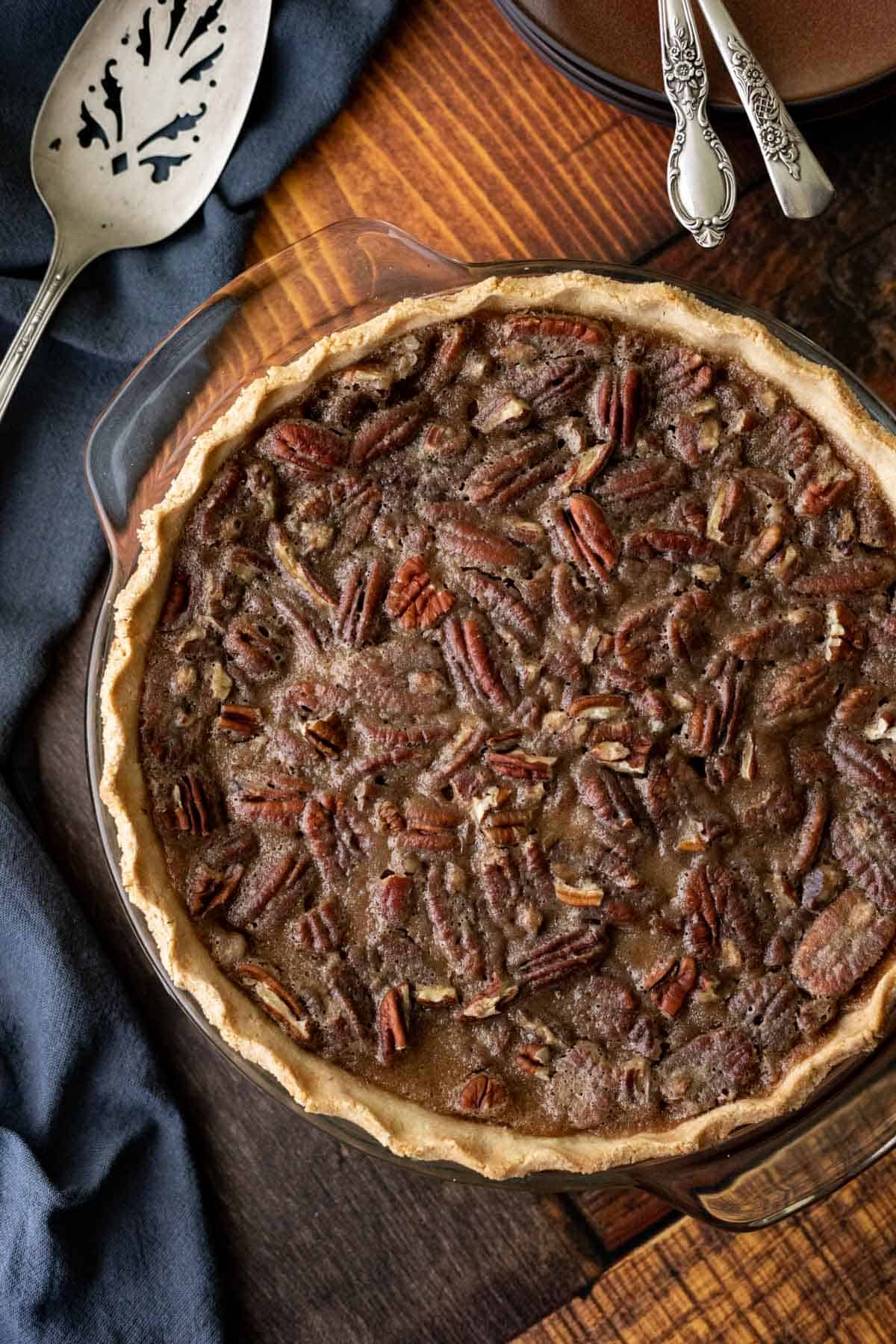 pecan pie in a glass pie plate on a dark wood table with a napkin and pie server on the left.