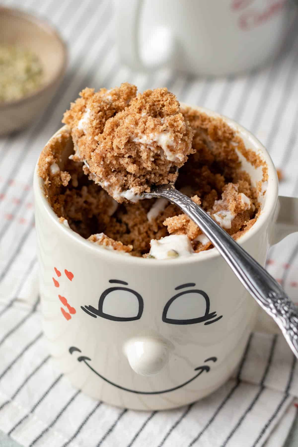 a spoonful of fluffy oat flour mug cake resting above a mug printed with a smiley face.