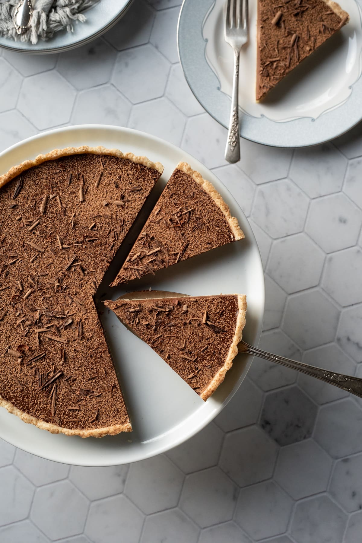 Dairy free dark chocolate tart on white cake stand with chocolate shavings on top.