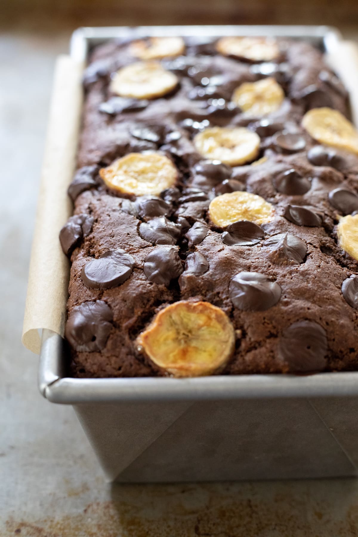 Loaf of chocolate banana bread in a pan, topped with sliced banana and chocolate chips.