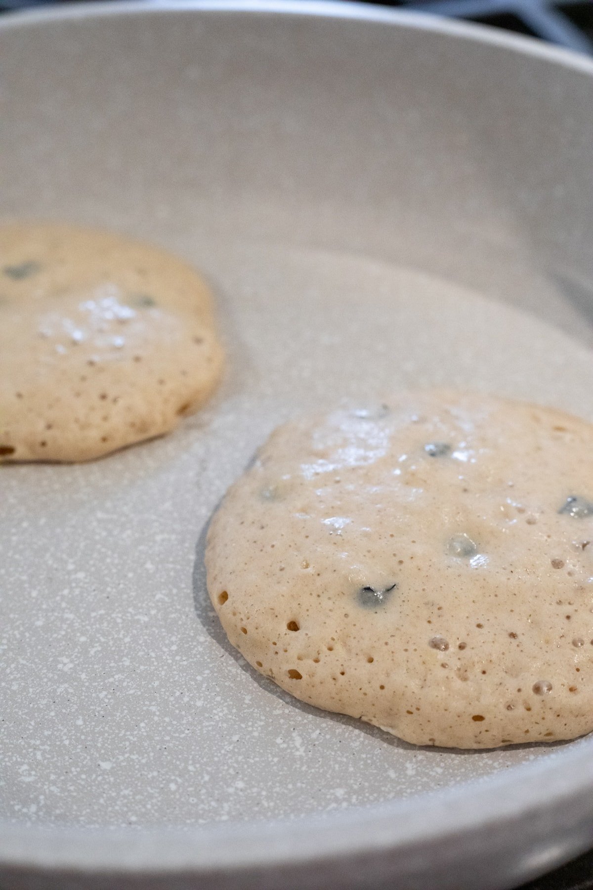 two blueberry pancakes cooking in a non-stick pan, showing bubbles.