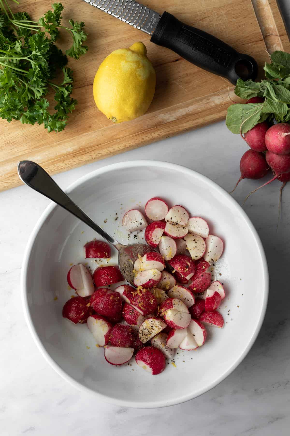 trimmed radishes in a large white bowl being tossed with oil, lemon zest, salt and pepper.