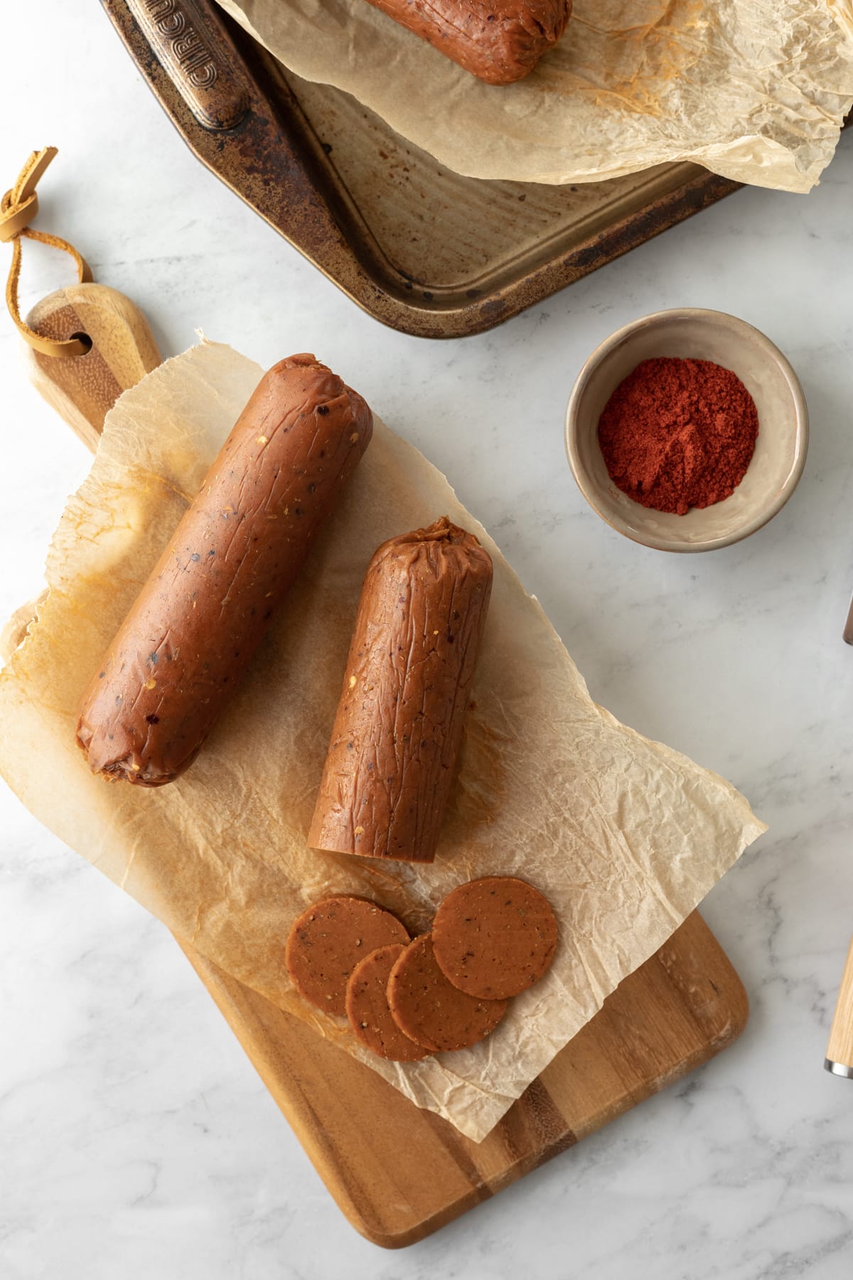 two logs of vegan pepperoni on a board with baking sheet and paprika in background.