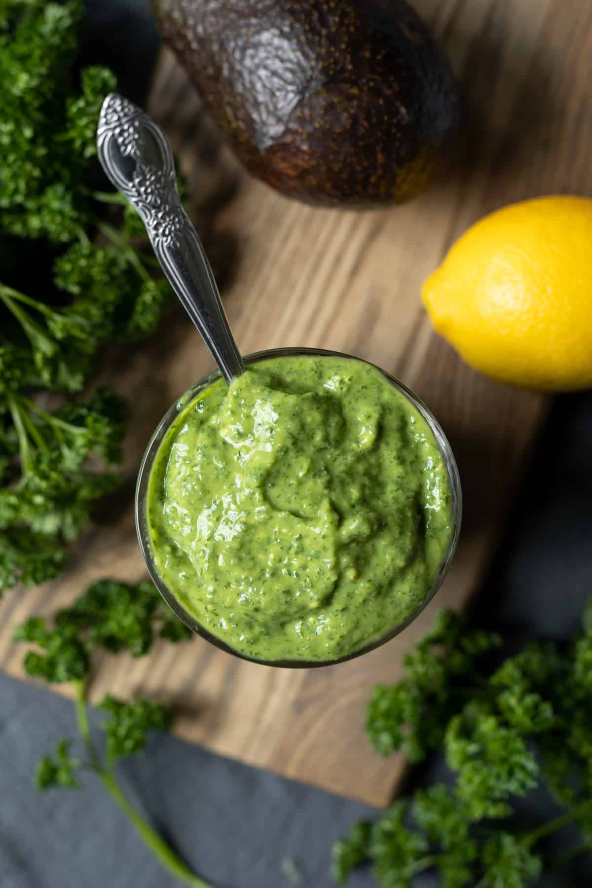 creamy green dressing in a glass surrounded by lemon, parsley, and a whole avodado