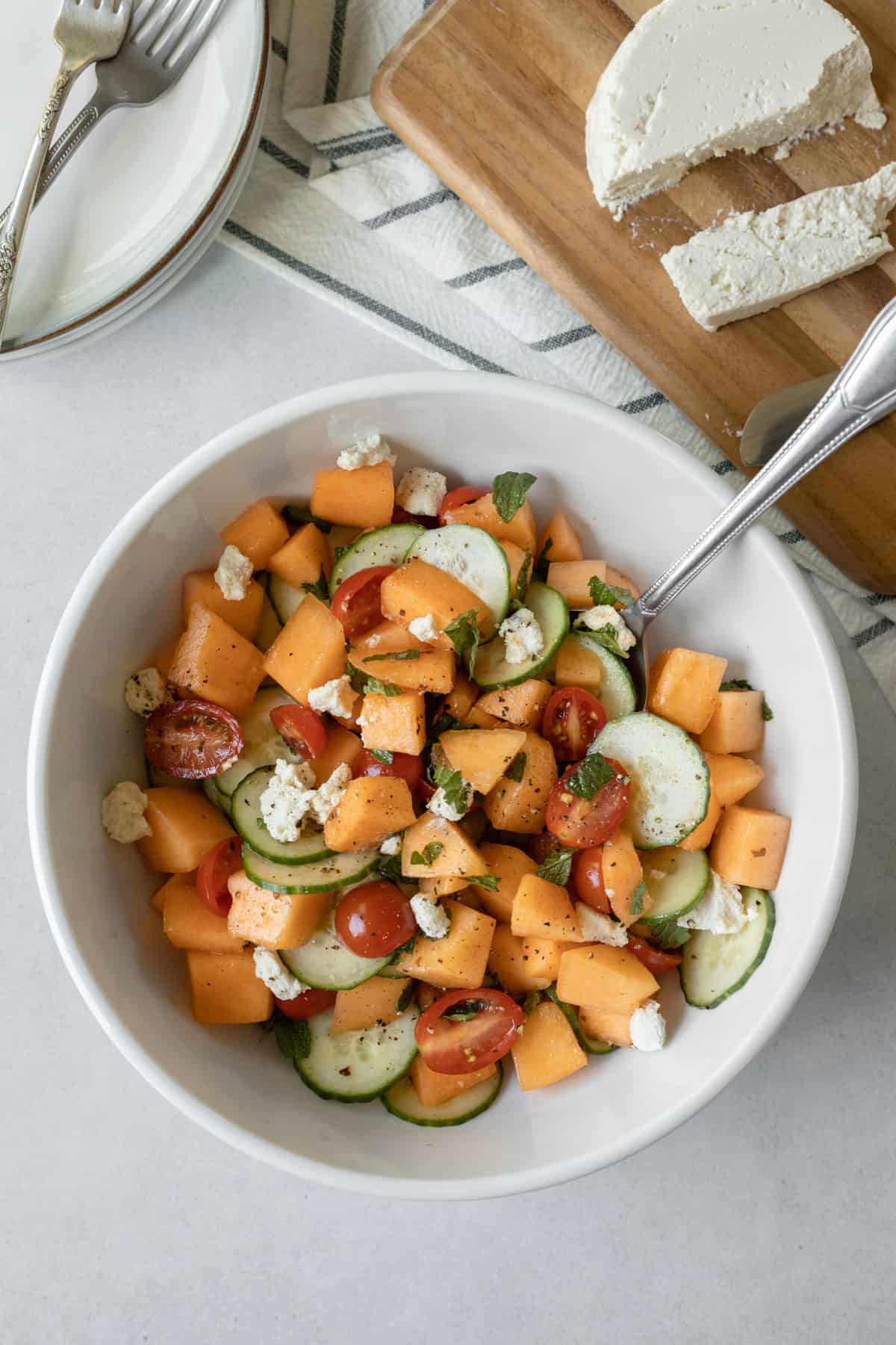 finished salad in a white bowl, vegan feta on a cheese board, and plates and utensils.