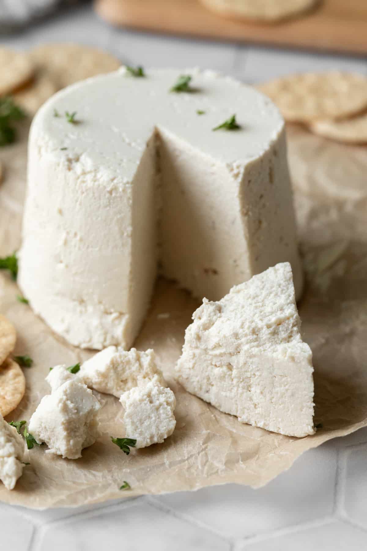 a wheel of vegan feta resting on parchment paper with some crumbled in front.