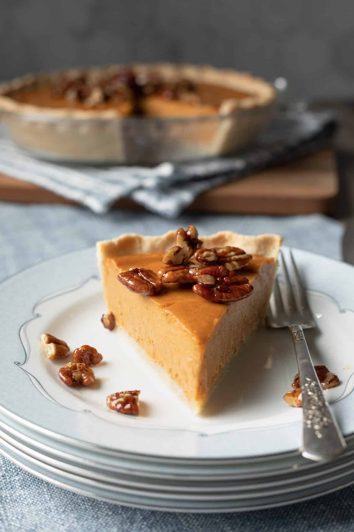 a slice of sweet potato pie topped with candied pecans with whole pie in background.