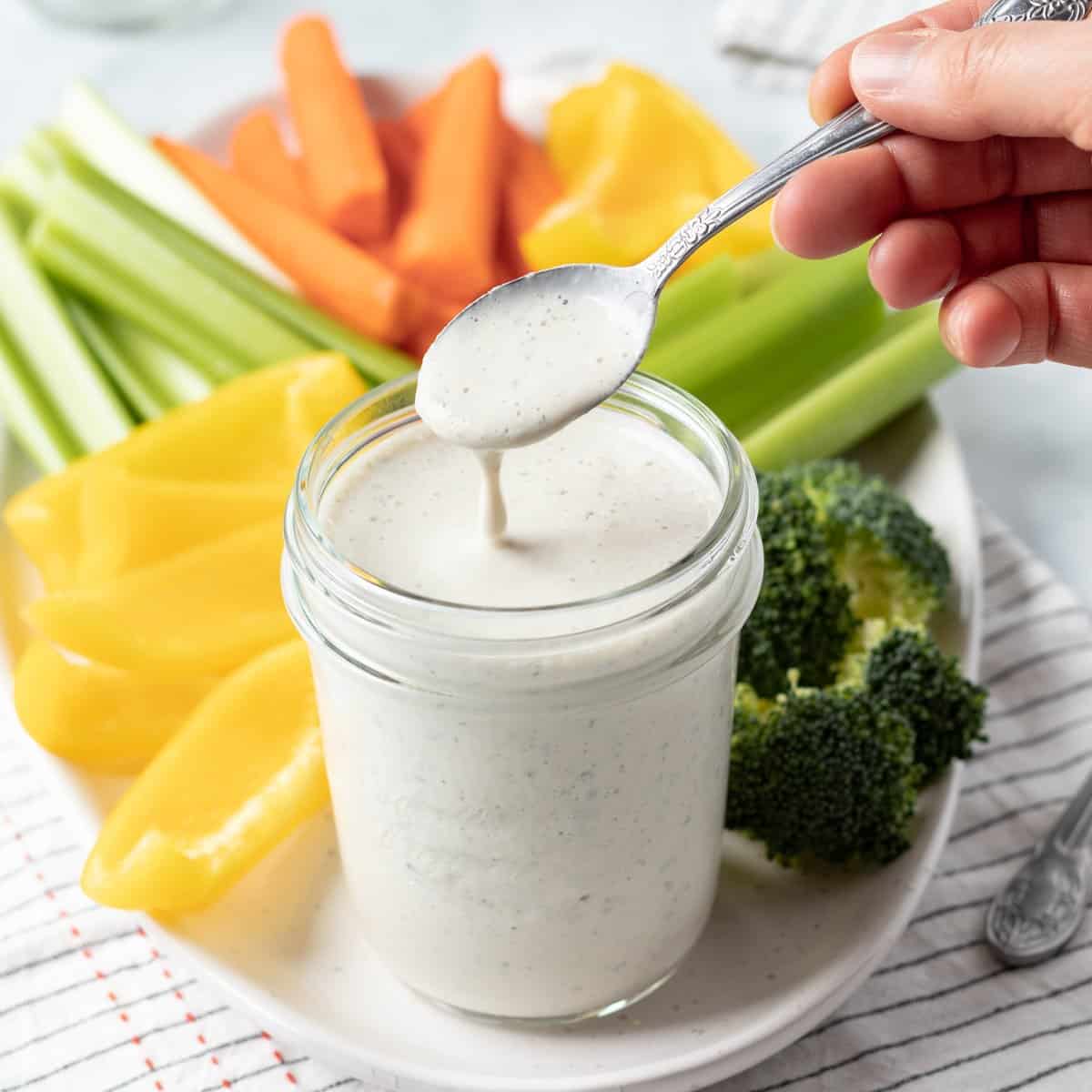 hand holding a spoon dripping ranch dressing over a jar.