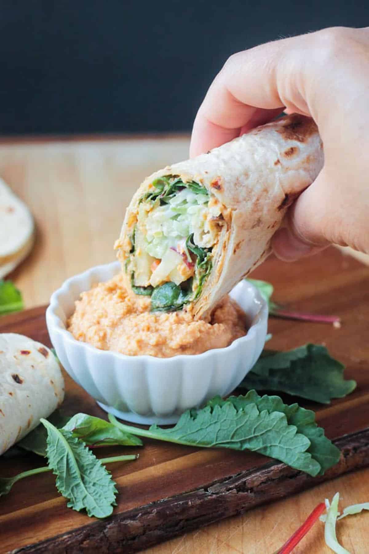 a hand dipping a veggie wrap into spicy hummus.