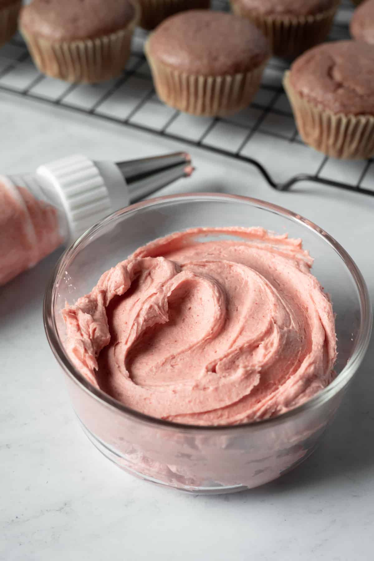 frosting in a small glass container with unfrosted strawberry cupcakes in background.