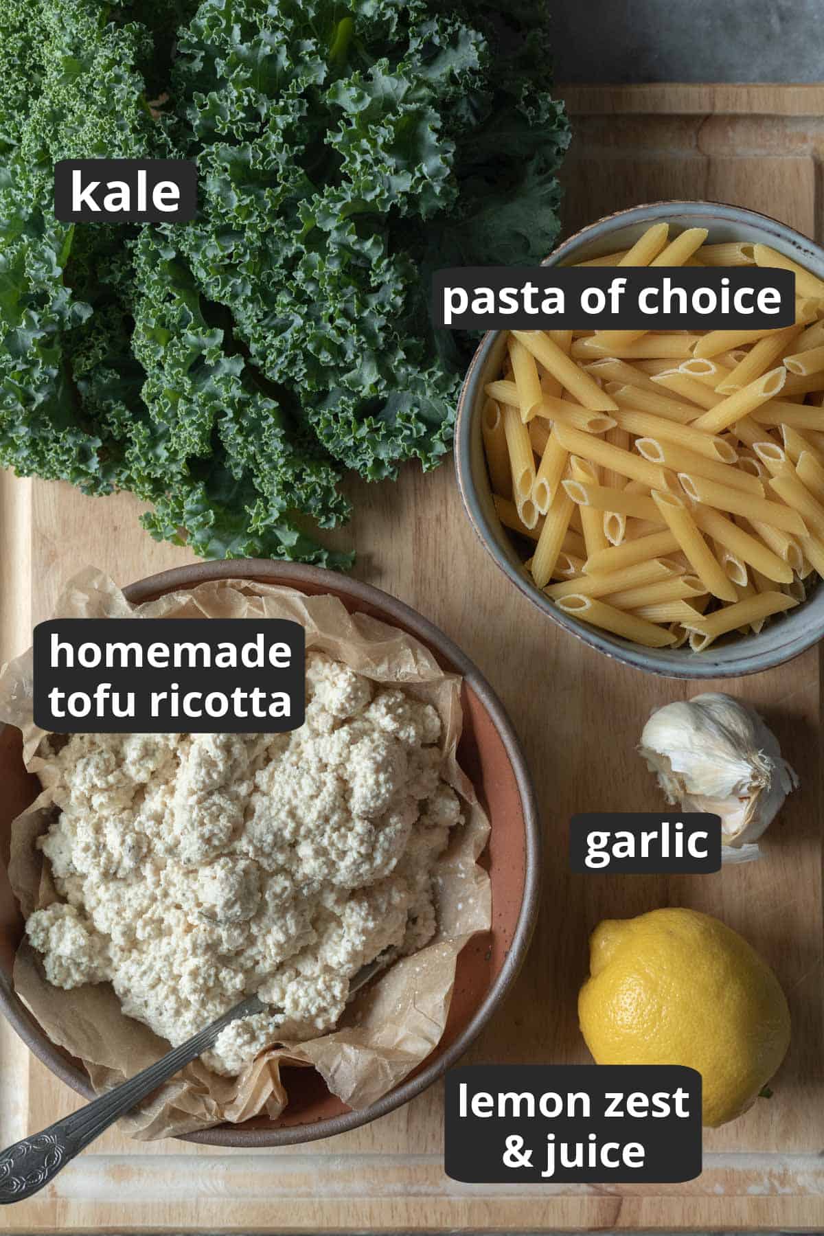 Ingredients on a cutting board: ricotta, kale, dry pasta, lemon and garlic.