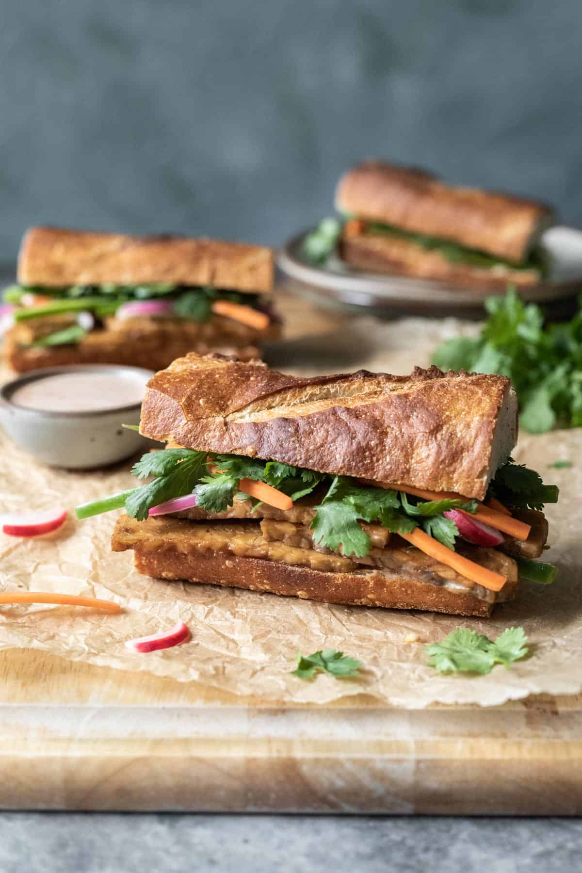 three tempeh banh mi sandwiches on a cutting board with spicy mayo nearby.