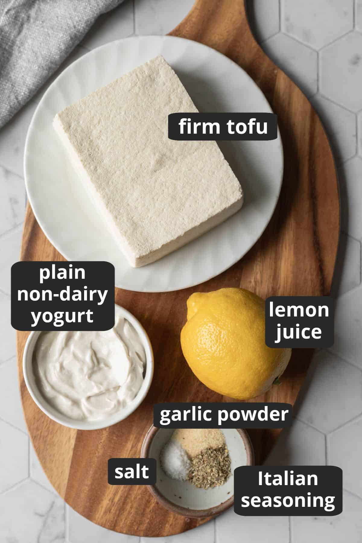 A labeled photo of ingredients needed for vegan ricotta: tofu, yogurt, lemon, salt, garlic, and herbs.