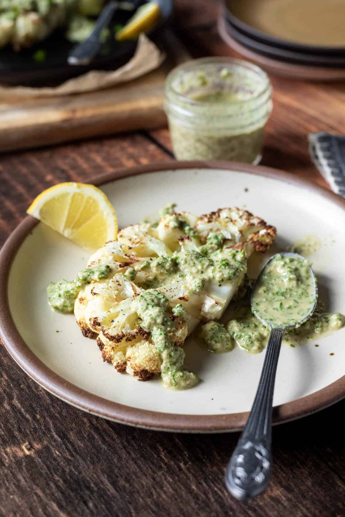 cauliflower steak on a cream colored plate with a spoon and nutty vegan parsley sauce.