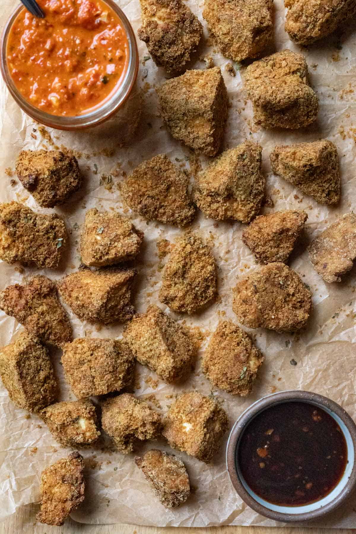 Batch of crispy tofu nuggets on parchment paper with 2 dipping sauces nearby.