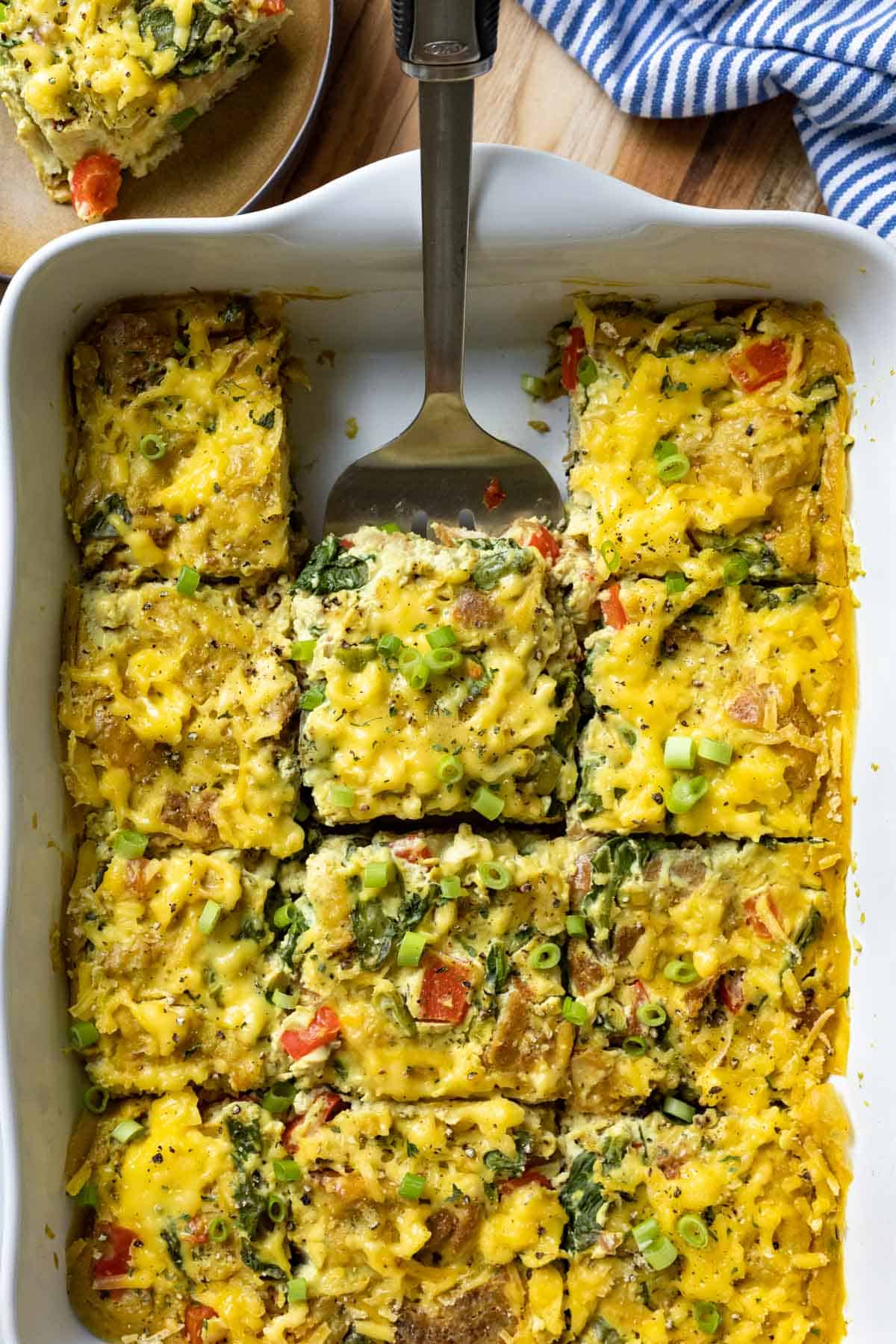 A spatula removing a serving of cheesy vegan breakfast casserole from a large white dish.