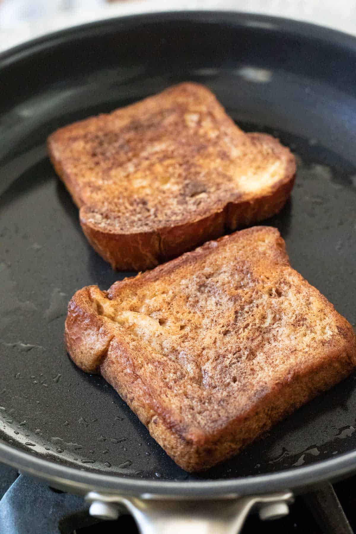 two thick slices of vegan French toast cooking in a non-stick pan.