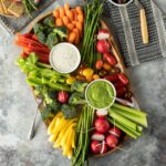 a colorful assortment of raw and blanched vegetables with two dips on a wooden tray.
