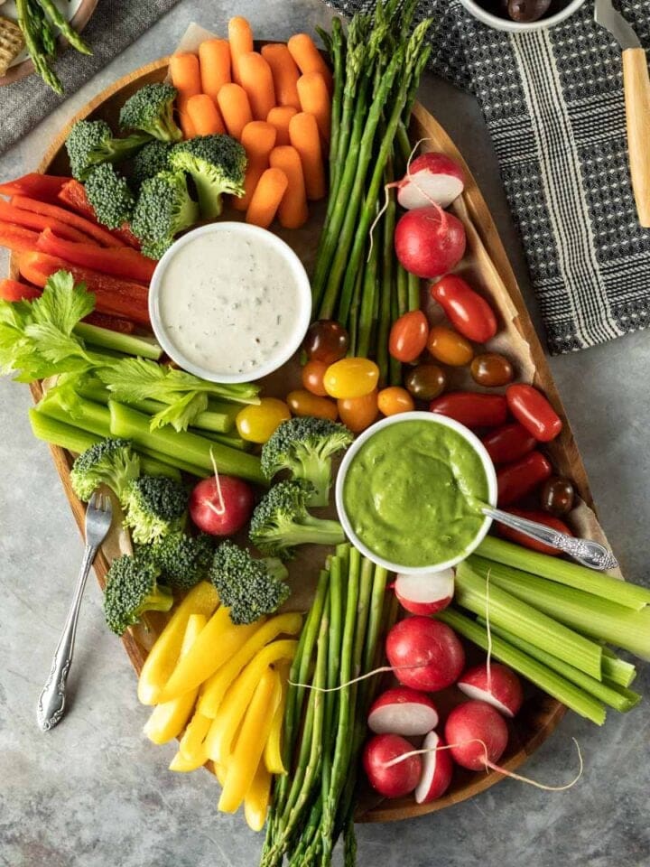 a colorful assortment of raw and blanched vegetables with two dips on a wooden tray.