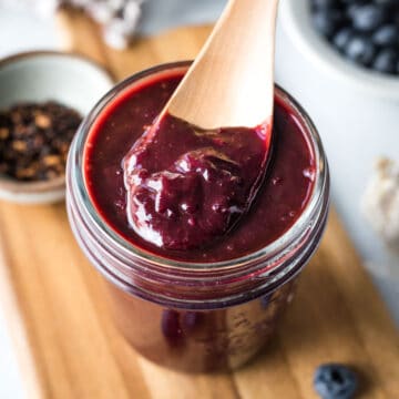 a jar of thick purple homemade barbecue sauce on a wood board with fresh blueberries nearby.