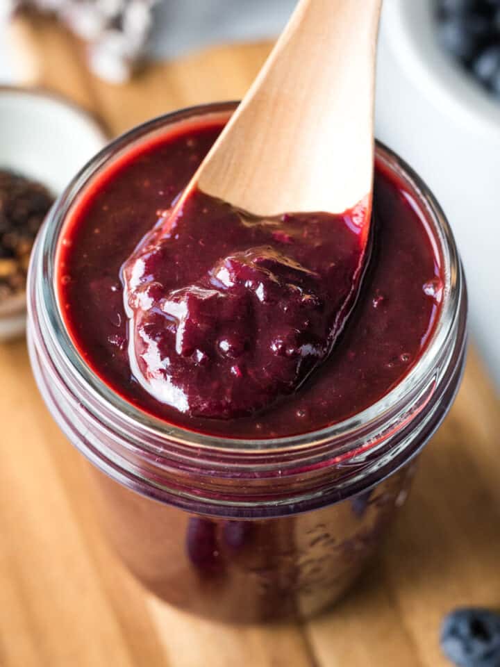 a jar of thick purple homemade barbecue sauce on a wood board with fresh blueberries nearby.
