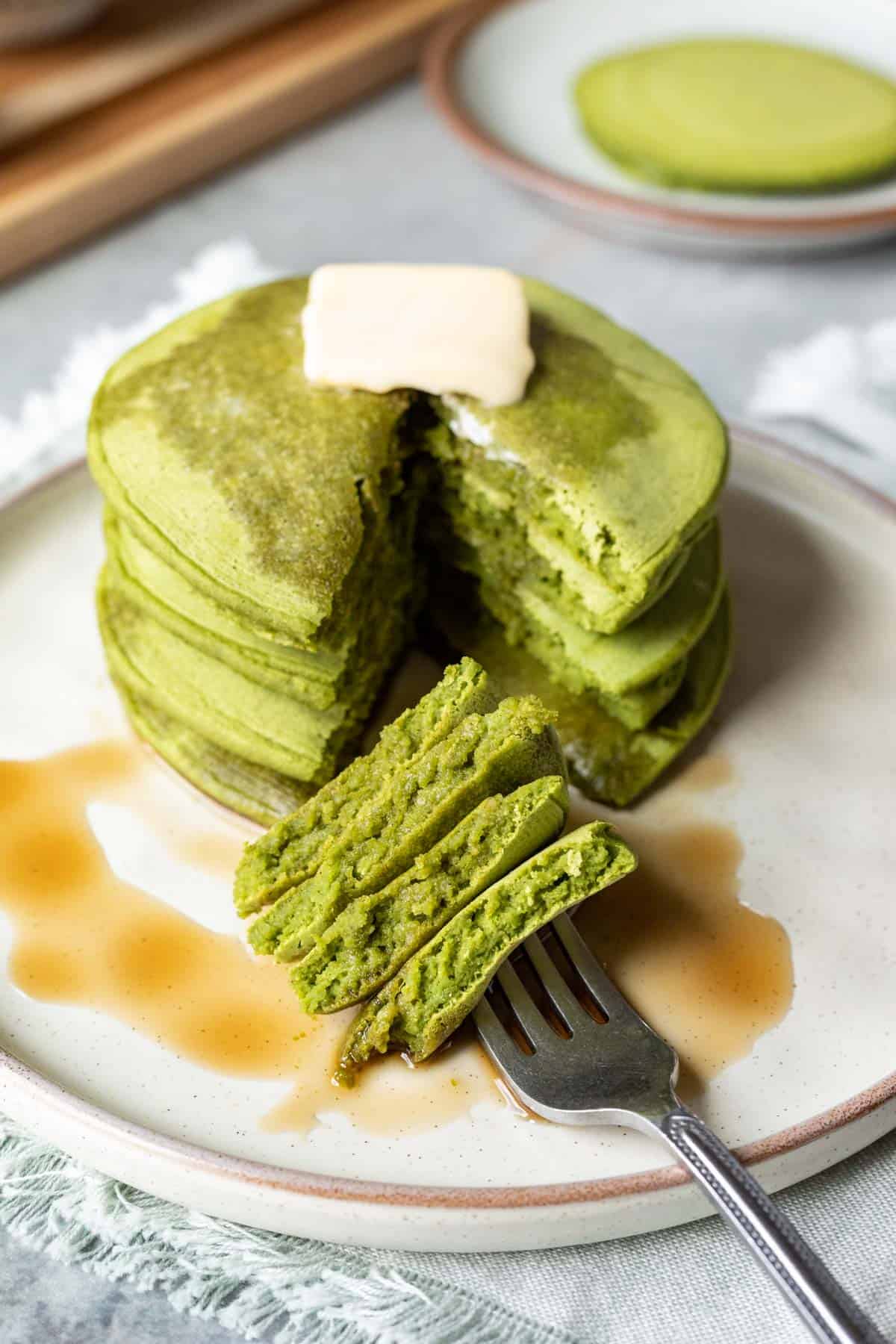 A bite of spinach pancakes on a fork with the full stack behind, drizzled with maple syrup and topped with a pat of butter.