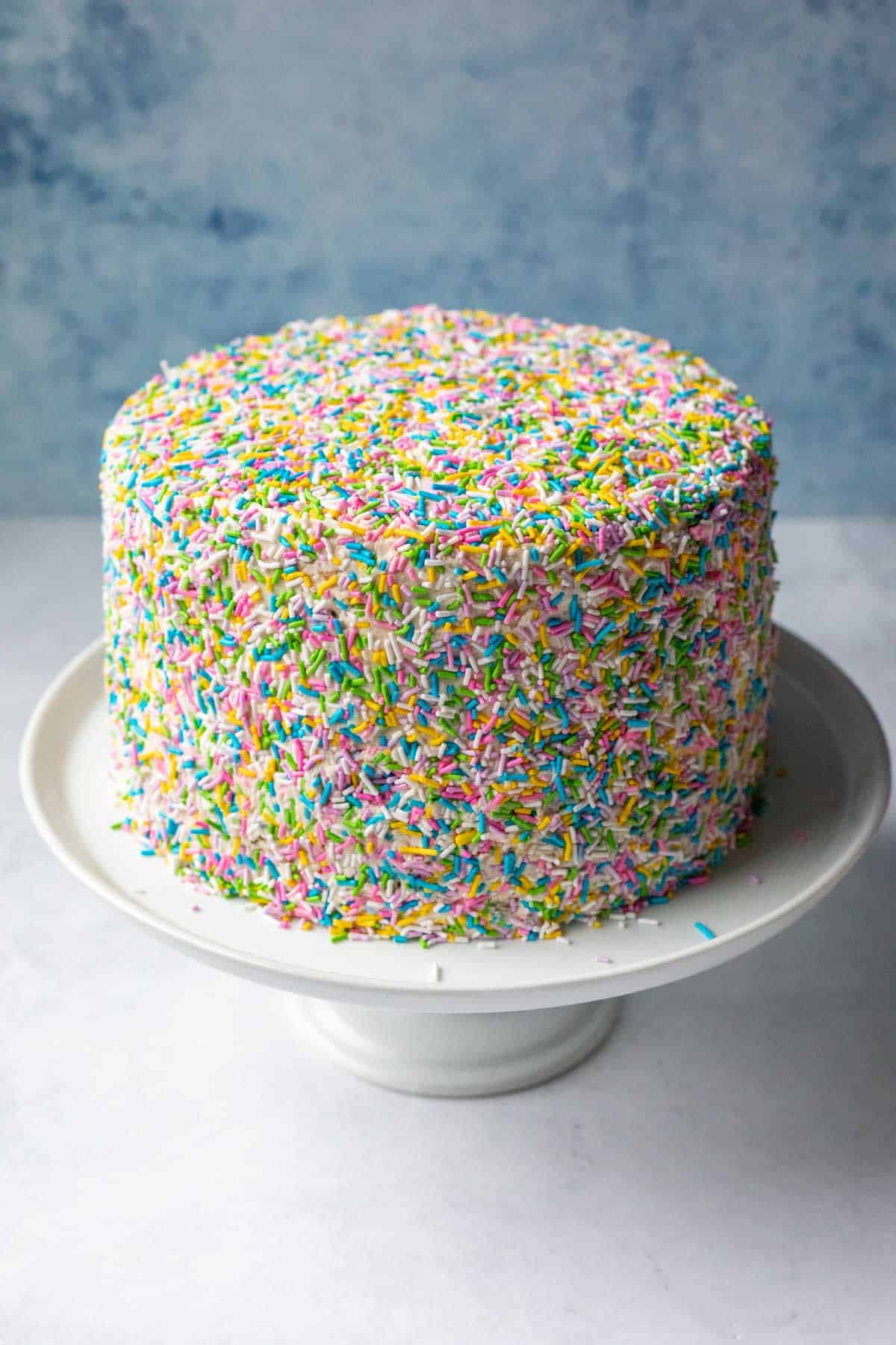 A 3-layer vegan vanilla cake coated in sprinkles resting on a white cake stand.