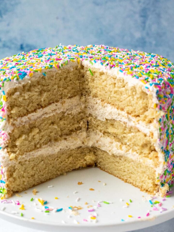 A 3 layer vegan vanilla cake decorated with sprinkles with pieces removed to show moist and fluffy texture.