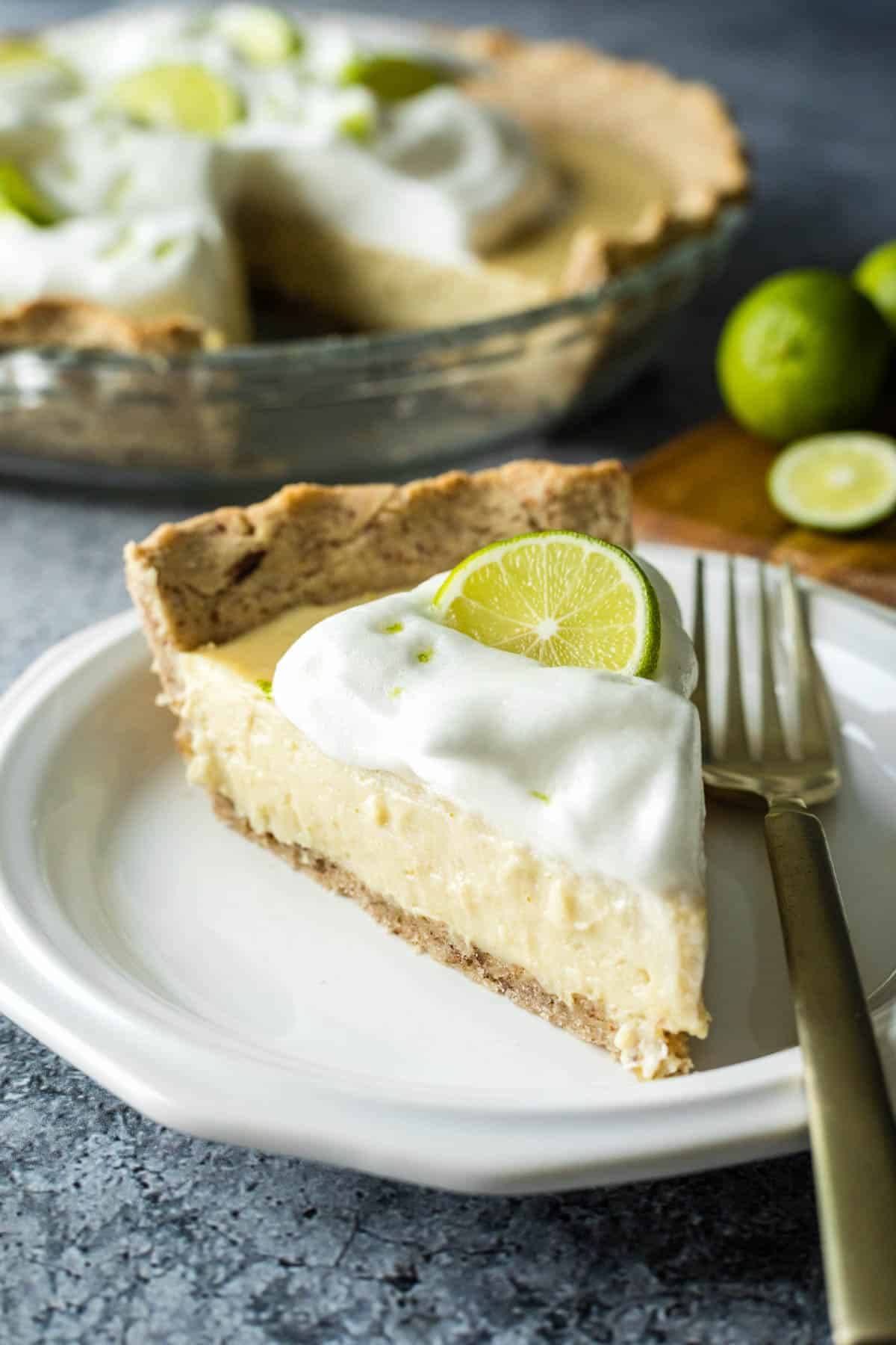 A slice of vegan key lime pie on a small white plate with the whole pie in the background.
