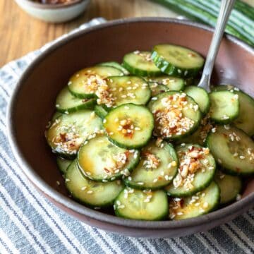 A dark red bowl filled with sliced cucumber tossed in spicy Asian dressing.