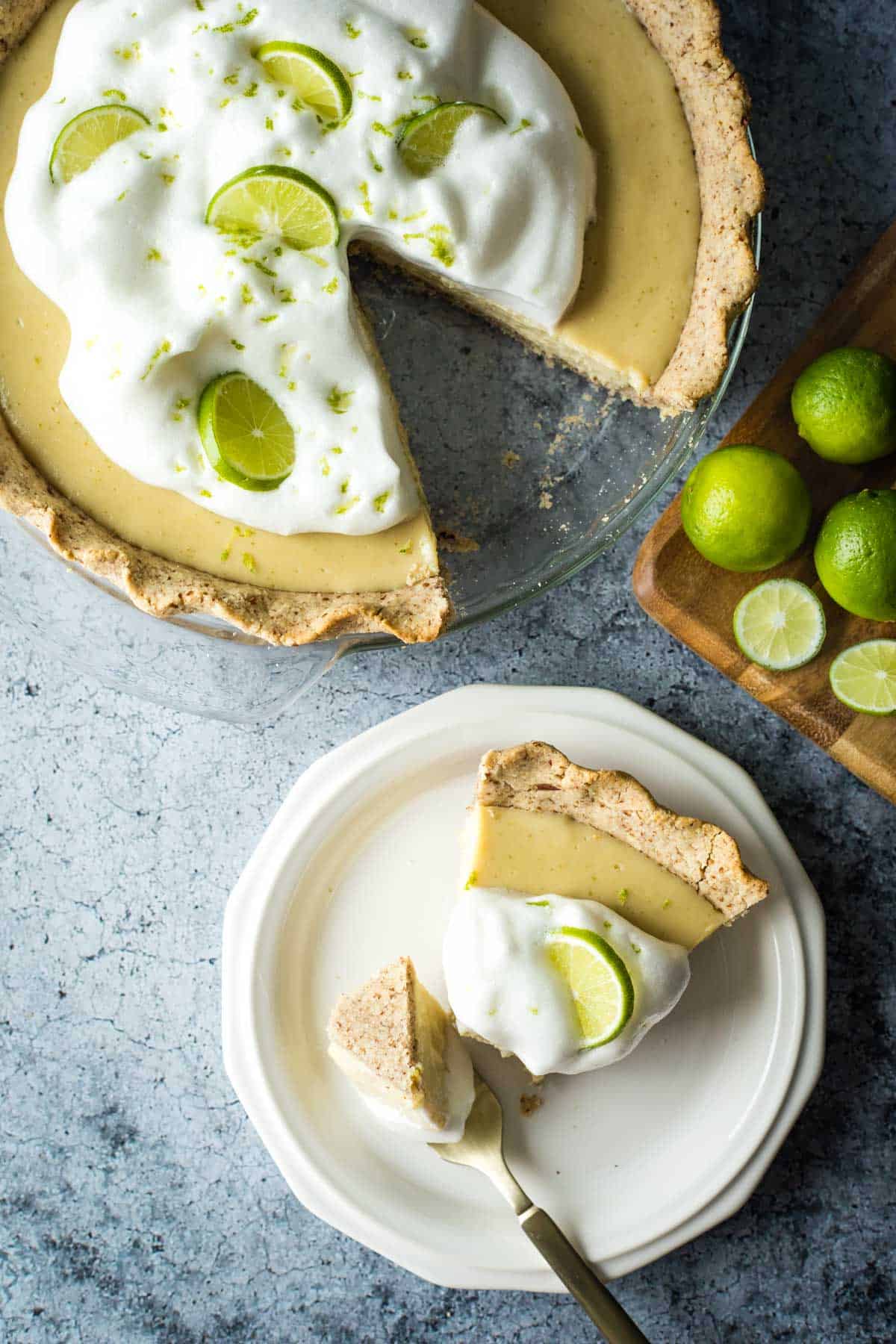 A slice of vegan key lime pie on a plate topped with aquafaba whipped cream and a slice of a key lime with the whole pie in background.