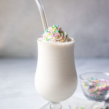 A vegan vanilla milkshake in a hurricane glass topped with non-dairy whipped cream and sprinkles.